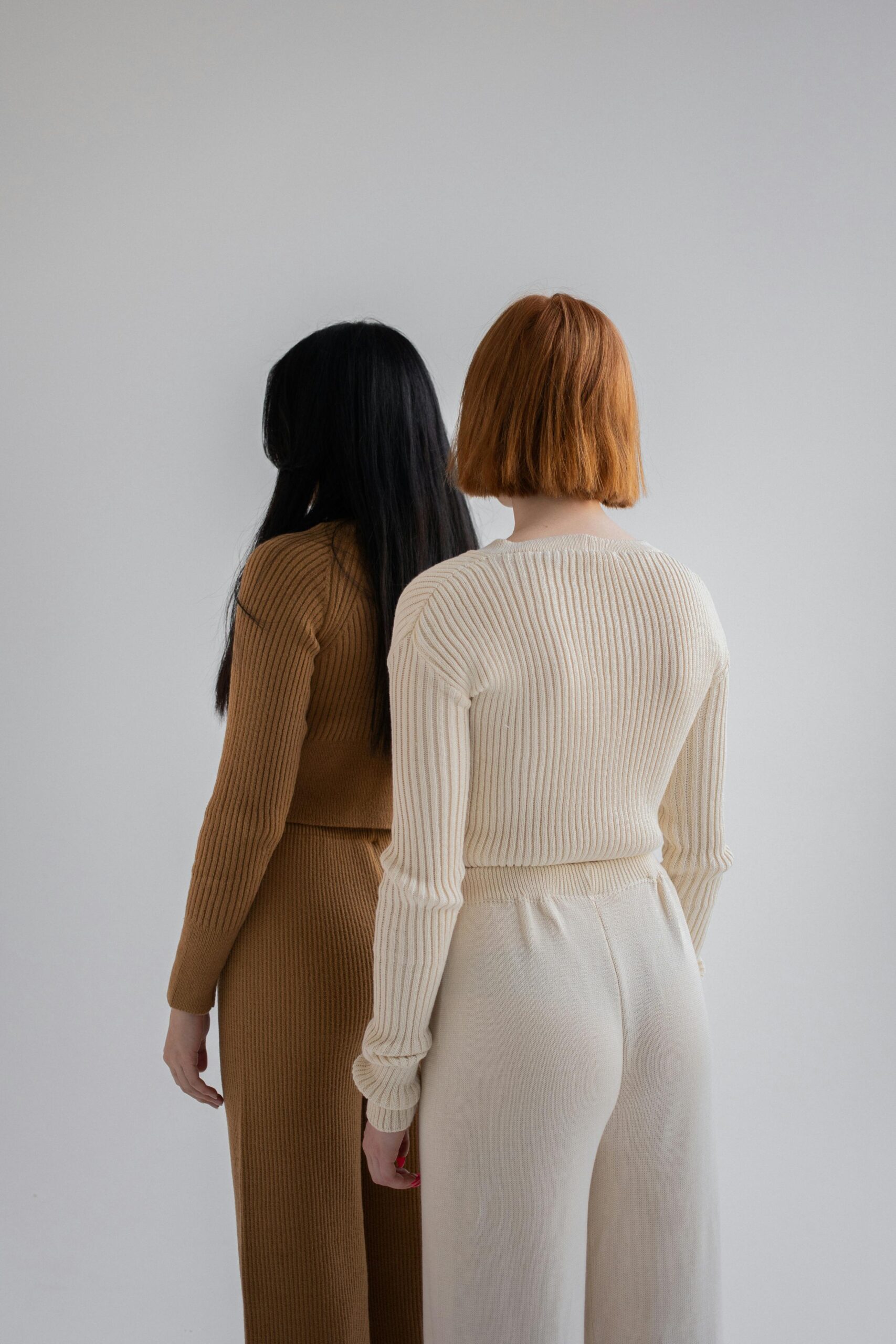Two women in knitted sweaters are seen from behind in a minimalist setting.